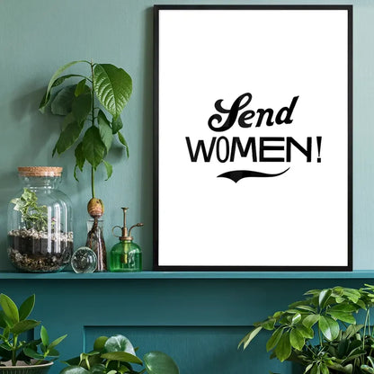 Black framed "Send Women!" typographic print on white archival paper, displayed on a teal shelf with plants.
