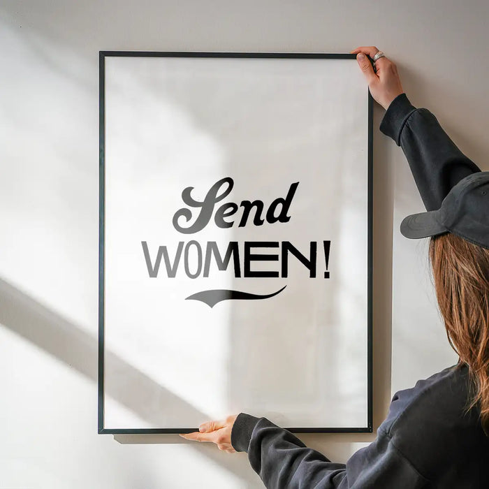 Black framed "Send Women!" typographic print on white archival paper, held by hands against a wall.