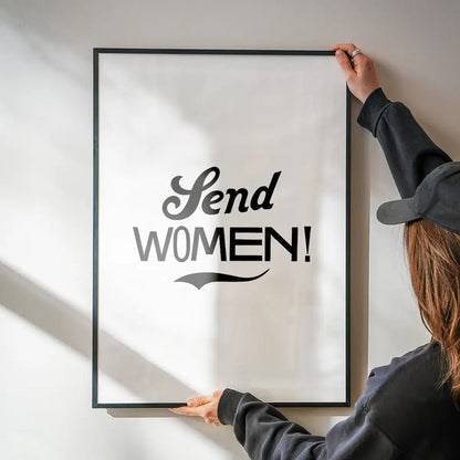 Black framed "Send Women!" typographic print on white archival paper, held by hands against a wall.