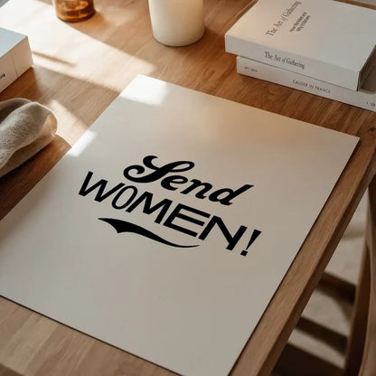 White archival art print with "Send WOMEN!" in bold black typography, on a wooden table.