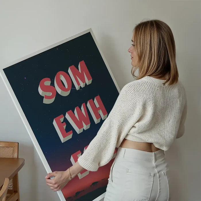 Woman holding a large "Somewhere" typography print with red 3D text on a dark blue starry sky and orange sunset.