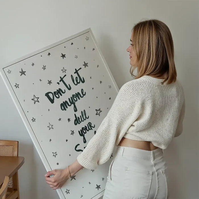 Woman holding a large white framed "Don't Let Anyone Dull Your Sparkle" typography print with stars.