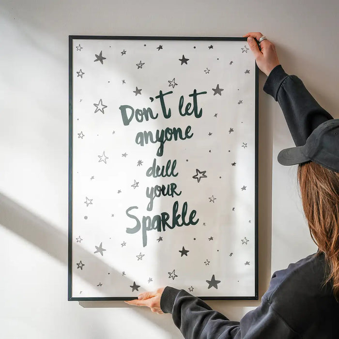 Black-framed "Don't Let Anyone Dull Your Sparkle" typography print with stars, held against a white wall.