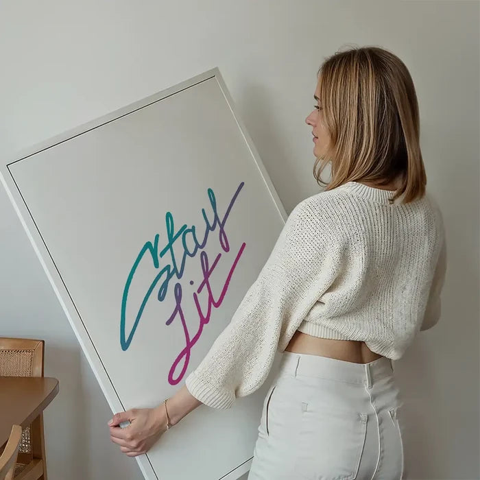 Woman holding a framed "Stay Lit" typographic print with a vibrant blue to pink gradient on white archival paper.
