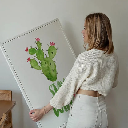 Woman holding a framed "Stay Wild" watercolor cactus print with pink flowers on a white background.