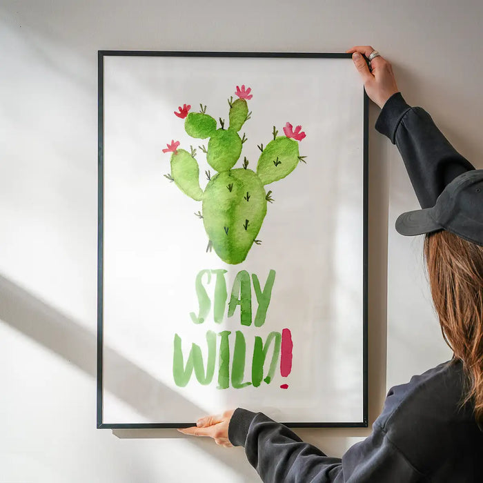 Watercolour cactus print with "Stay Wild!" text in a black frame, held by hands on a white wall.