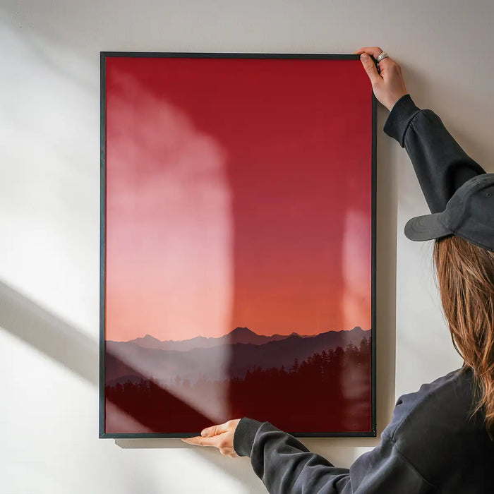 Person hanging a framed duotone red and orange sunset print with dark mountains and trees on a white wall.