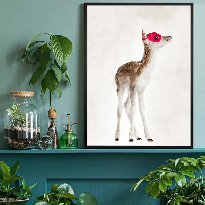 Watercolor print of a fawn in a red superhero mask, framed in black, on a teal shelf with plants.