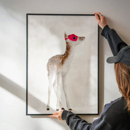 Watercolor fawn superhero print with a red mask in a black frame, held by hands on a white wall.