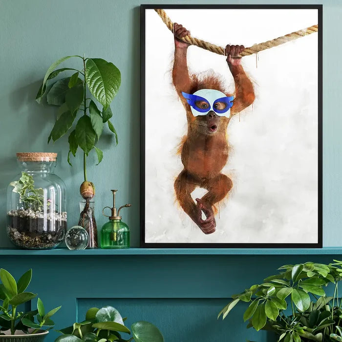 Watercolor orangutan hero print with blue mask swinging on a rope, framed in black, on a green shelf.