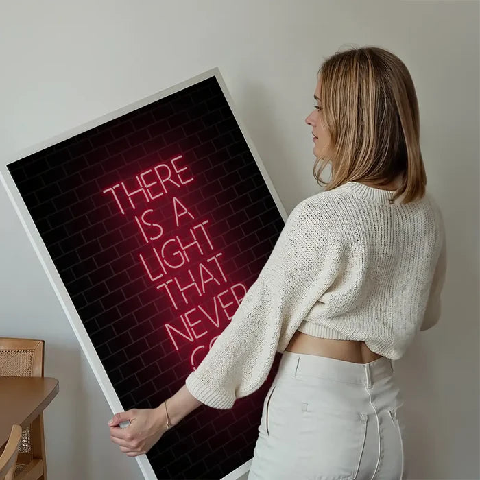 Woman holding a framed art print with "THERE IS A LIGHT THAT NEVER GOES OUT" in red neon-style text on a brick background.