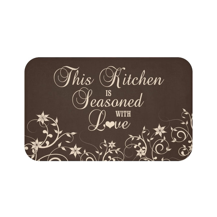 Floor mat with brown background and tan vine pattern across bottom of the mat. The phrase "This Kitchen is Seasoned with Love" is written in tan cursive script in the center.