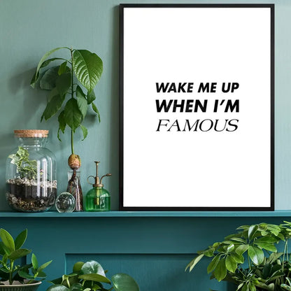 Black framed "Wake Me Up When I'm Famous" quote print on a white background, displayed on a green shelf with plants.