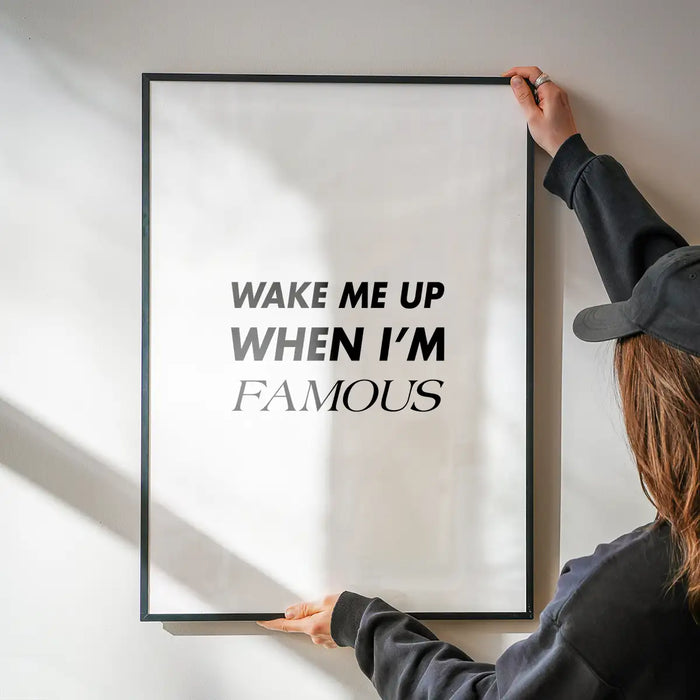 Black framed "Wake Me Up When I'm Famous" typographic print on white archival art paper, held by hands.