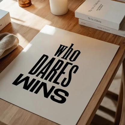 White typographic print with "WHO DARES WINS" in bold black text on a wooden table.