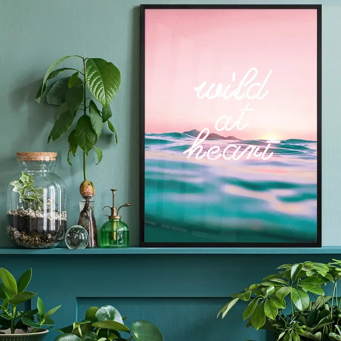 Neon "wild at heart" ocean print with pink sunset and turquoise water in a black frame, on a teal shelf.