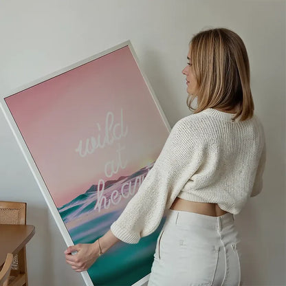 Woman holding a framed "Wild At Heart" neon print with pink sky and ocean waves.