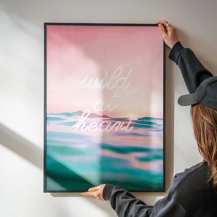 Framed "Wild At Heart" neon print with pink sky and teal ocean, held by hands on a white wall.