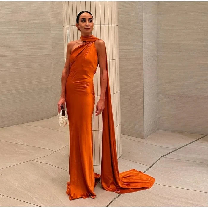 Woman in an orange evening gown with a long train standing against a neutral wall.