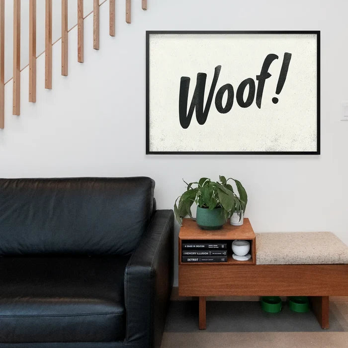 Black framed "Woof!" brush script typography print on a textured cream background, hanging above a modern wooden bench.