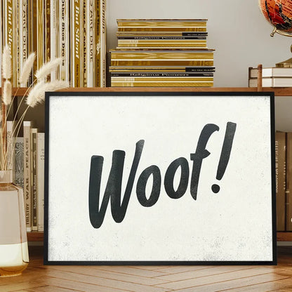 Black framed "Woof!" brush script typography print on a textured white background, displayed on a wooden shelf.
