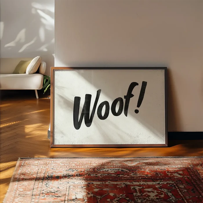 Minimalist "Woof!" brush script typography print in a dark wood frame, leaning on a wall above a patterned rug.